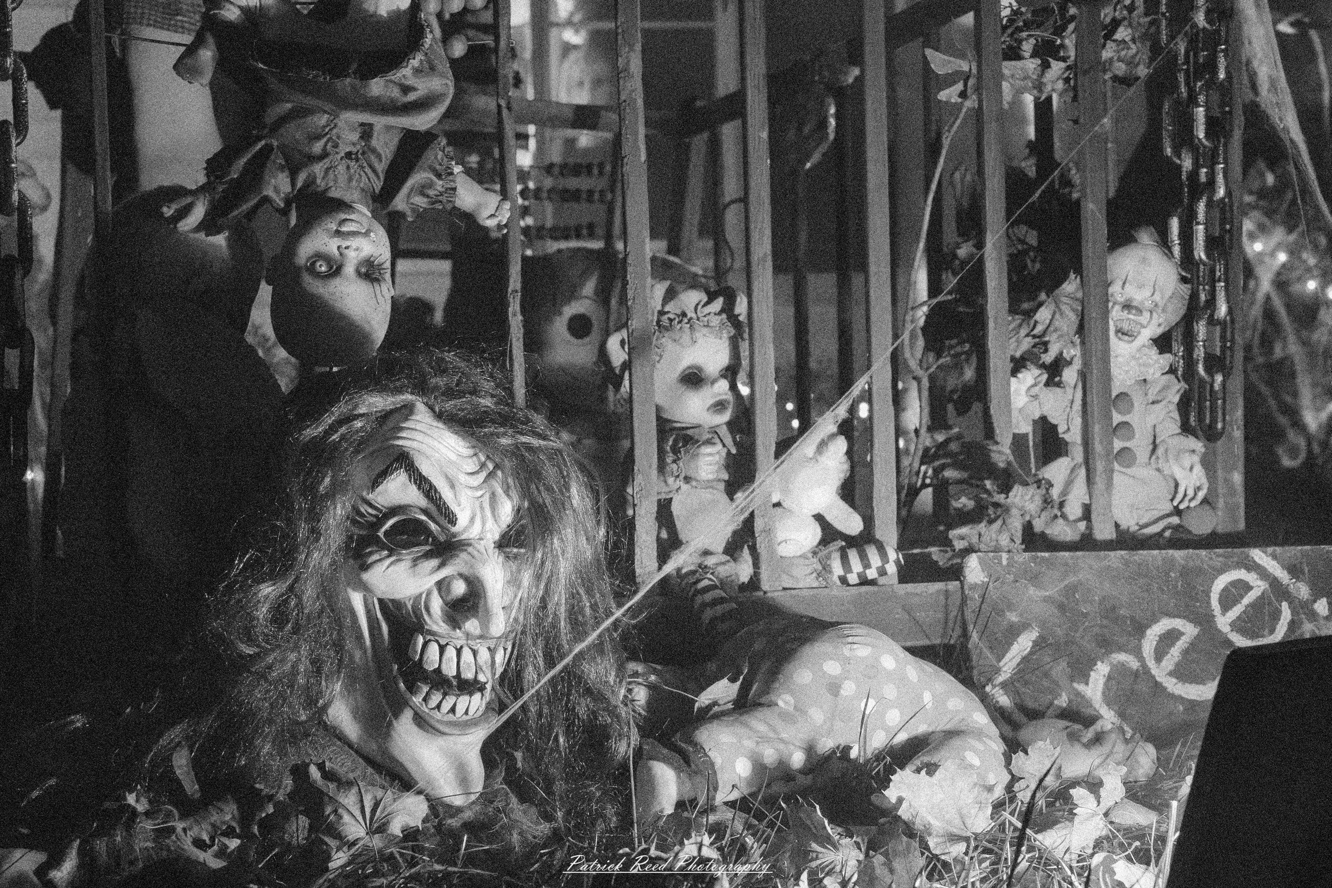 A chilling scene of a cluttered shelf filled with old dolls, each with cracked porcelain faces and empty, staring eyes. In the center, a sinister clown mask with exaggerated features and hollow eyes sits menacingly among the dolls. The black-and-white color scheme enhances the eerie atmosphere, casting deep shadows that add to the sense of unease. Dust settles on the dolls, and the dim lighting emphasizes their worn, aged appearance. The entire image exudes an unsettling stillness, as if the dolls and mask might come to life at any moment.